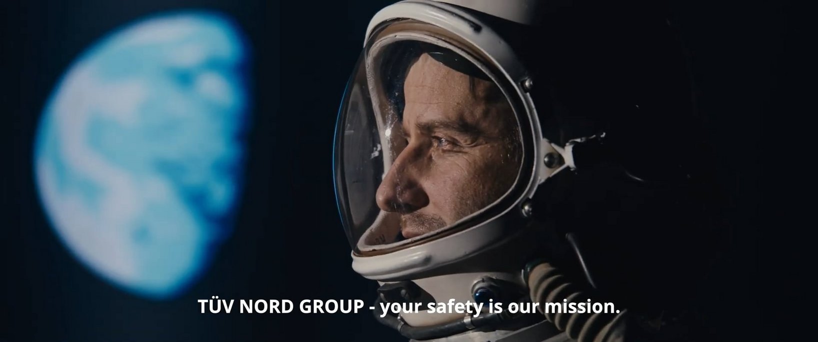 Das Bild zeigt eine Nahaufnahme eines Astronauten mit einem weißen Raumhelm und reflektierendem Visier. Er blickt mit einem zufriedenen und nachdenklichen Gesichtsausdruck in die Ferne, während im Hintergrund die Erde verschwommen zu sehen ist. Die Szene ist in dunklen Farben gehalten, was den Kontrast zwischen dem Astronauten und dem Planeten verstärkt. Am unteren Bildrand ist der Schriftzug ‚TÜV NORD GROUP – Ihre Sicherheit ist unser Antrieb‘ eingeblendet. Dieses Bild ist das Thumbnail für den Imagefilm der TÜV NORD GROUP und symbolisiert Fortschritt, Innovation und Sicherheit.